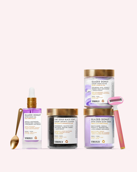 The Glazed Donut Shave Set from Truly Beauty includes After Shave Glow Cream, Soft Serve Whipped Shave Butter, a serum with dropper, black soap jar, gold razor, and gold spoon—all displayed against a pale pink background.