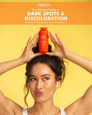 A woman with curly hair holds an orange bottle above her head, puckering her lips. Above her, text reads: The Ultimate Guide to Dark Spots & Discoloration—discover Laser Spot Dark Spot Treatment E-Book by Truly Beauty.