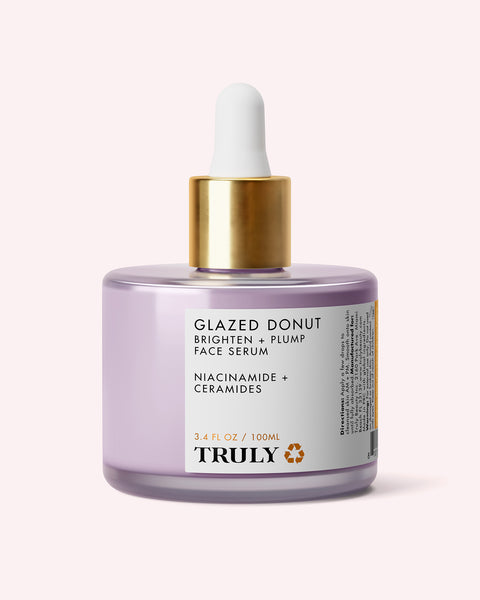 A lavender glass bottle of Truly Beauty Glazed Donut Brighten + Plump Face Serum, featuring a gold and white dropper cap. The label showcases its hydrating formula with powerhouse ingredients like niacinamide and ceramides, and reads “BRIGHTEN + PLUMP.” Perfect for achieving Glazed Donut Skin, this bottle contains 3.4 fl oz (100 ml) of serum.