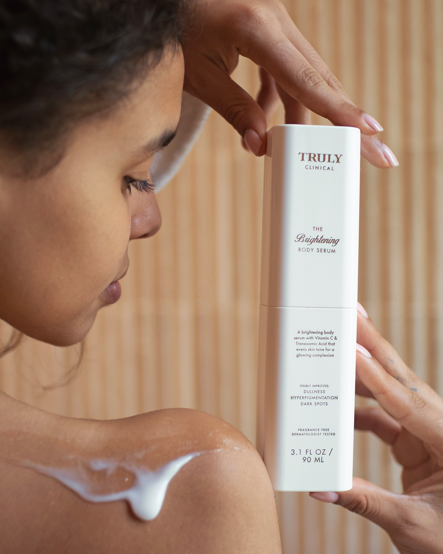A woman applies a white lotion to her shoulder while holding a bottle of The Body Treatments Complete Collection by Truly Beauty, showcasing effective hydrating body care against a neutral background.