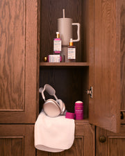 A wooden cabinet with an open door displays Truly Beauty’s The Viral Edit skincare set, a large tumbler with a straw, purple headphones on a white towel, and a pink container of wellness powder.