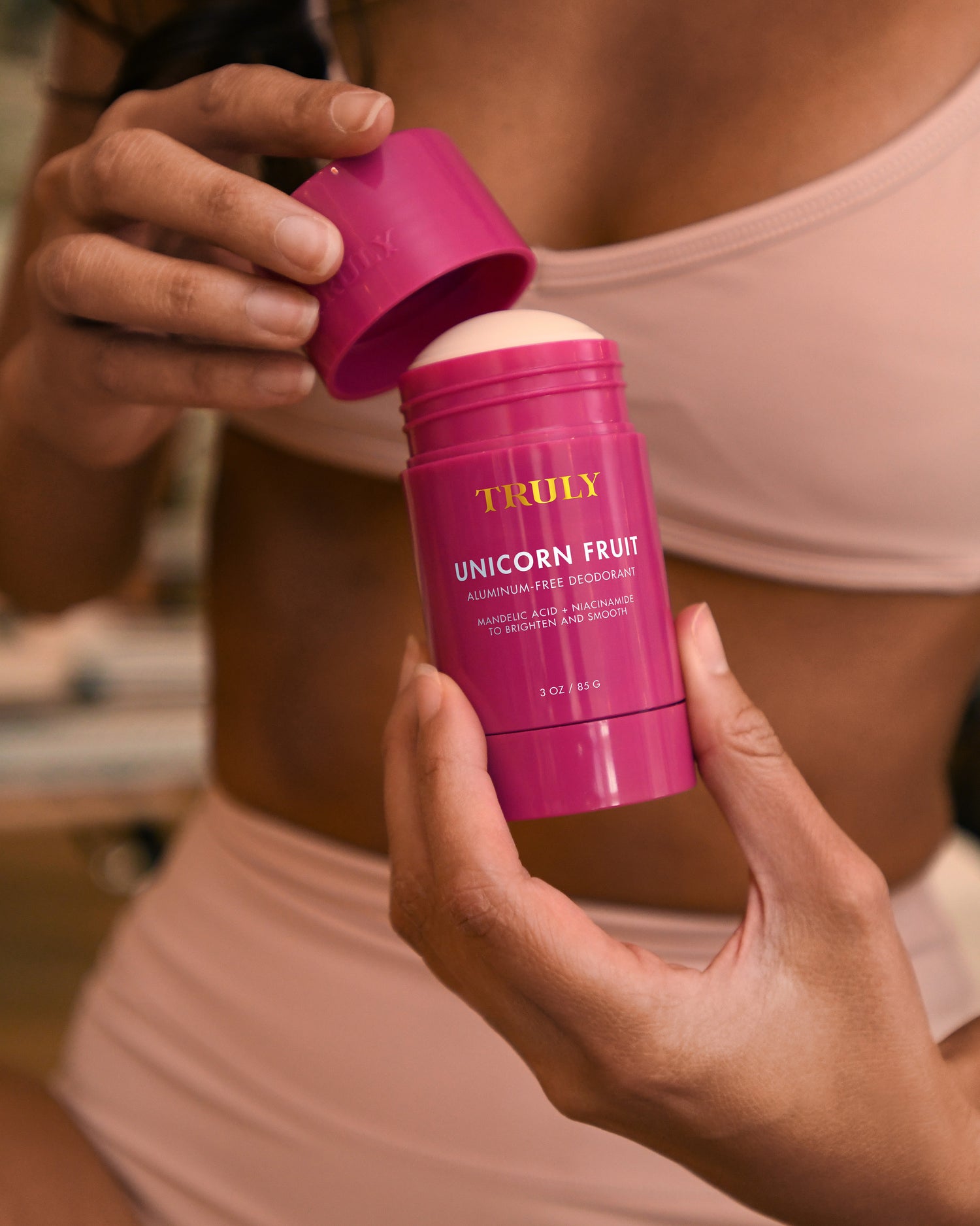A person in a beige bikini opens a pink stick of Truly Beauty’s The Viral Edit deodorant, holding it close to their body and lifting the lid with the other hand.