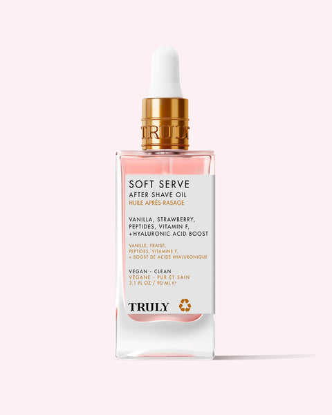A glass dropper bottle of Soft Serve After Shave Oil features a gold cap, clear pink liquid, and a white label listing ingredients like vanilla, strawberry, peptides, vitamin F, and hydrating hyaluronic acid.