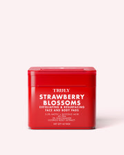 A red container labeled Truly Beauty Strawberry Blossom Exfoliating & Resurfacing Face & Body Pads, featuring product details in white text, sits on a light pink background. These pads utilize lactic and glycolic acids for gentle yet effective exfoliation.