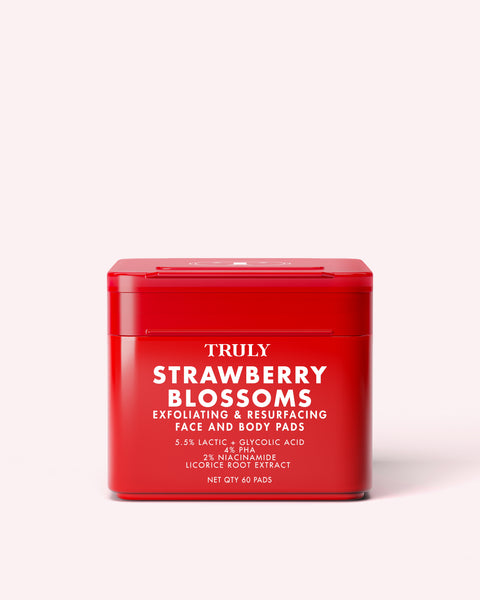 A red container labeled Truly Beauty Strawberry Blossom Exfoliating & Resurfacing Face & Body Pads, featuring product details in white text, sits on a light pink background. These pads utilize lactic and glycolic acids for gentle yet effective exfoliation.