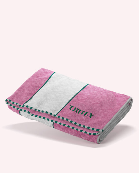 The Signature Terry Oversized Towel by Truly Beauty features pink and white stripes, green edging, and “TRULY” embroidered in green on the white stripe. Made from durable terry cloth, it’s shown folded on a light pink background.