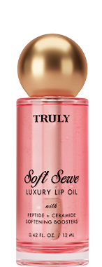A bottle of Truly Lip Oil Soft Serve with a gold cap sits before a pink swoosh, strawberry, and ice cream cone. The label features product details.