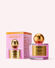 A 50 ml bottle of Truly Beauty Unicorn Fruit Eau De Parfum, featuring a gold label and round gold cap, is displayed beside its pink and orange box with gold lettering. This fragrance has a fruity, feminine scent inspired by candy floss.