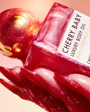 A close-up of Truly Beauty’s Cherry Baby Luxury Body Oil—a pink bottle with a gold cap, adorned with glossy cherry-red drips against a soft pink background—delivers glowing hydration and radiant shimmer.