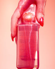 A hand with shiny, pink glittery nails holds a glossy bottle of Truly Beauty’s Cherry Baby Luxury Body Oil against a pink backdrop. The bottle and hand both glisten with matching shimmer, echoing the product’s radiant, sparkling appeal.