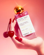 A hand with long red nails holds a pink bottle of Truly Beauty Cherry Baby Luxury Body Oil, featuring radiant shimmer, against a pink gradient background; the fingers also hold two cherries by their stems.