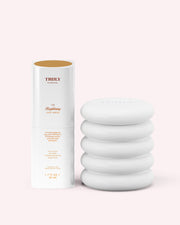 A cylindrical bottle labeled “Truly Beauty The Brightening Face Routine” stands open, revealing a gold inner lid, beside a white, stacked ceramic object on a pale pink background.