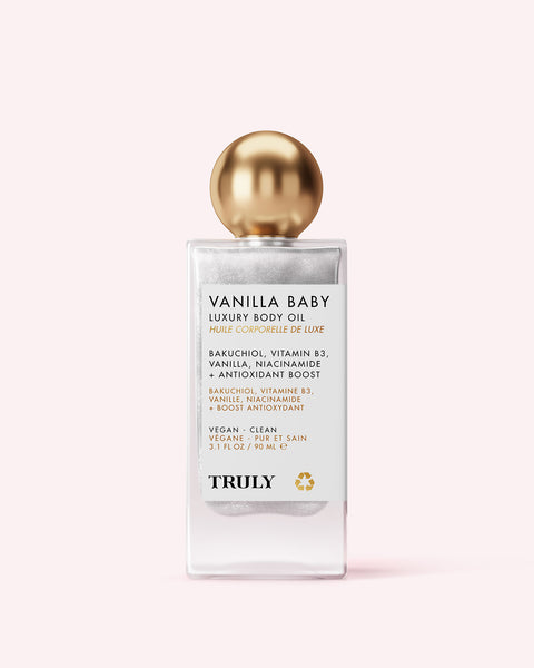 A clear glass bottle of Truly Beauty Vanilla Baby Luxury Body Oil with a gold round cap and white label listing tightening, anti-aging ingredients like bakuchiol, vitamin B3, and niacinamide, set against a light pink background.