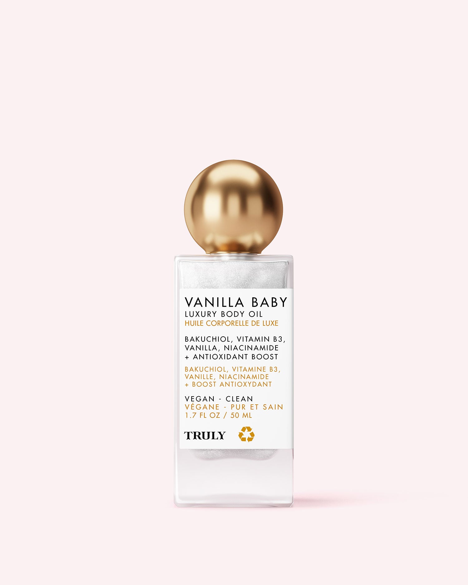 A bottle of Truly Beauty Vanilla Baby Luxury Body Oil with a gold spherical cap and white label, featuring black and gold text, sits against a pale pink background. The label touts plumping, anti-aging benefits, key ingredients, and product features.