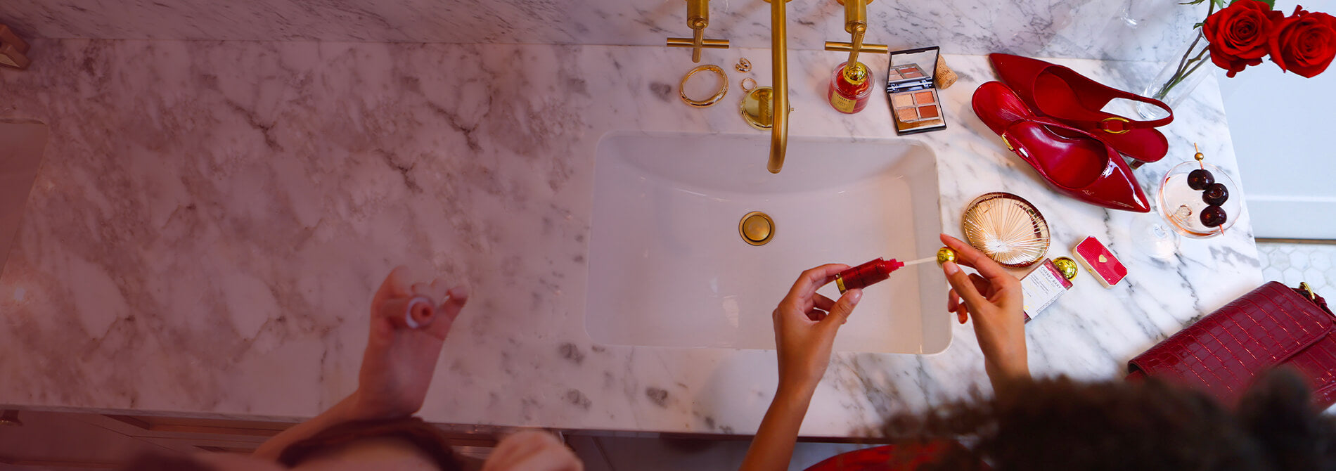 Someone holds a Truly Lip Oil over a marble bathroom sink, surrounded by red roses, red heels, makeup, jewellery, and cherries, with gold faucet and accessories in view.