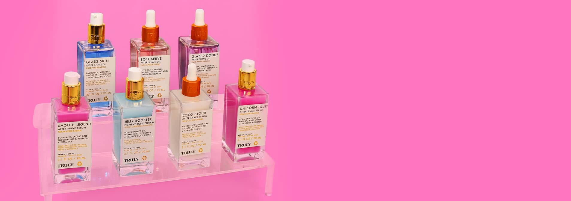 A set of cosmetic bottles in a clear acrylic stand, featuring various shades of pink, blue, and peach liquid with gold caps, set against a solid bright pink background.