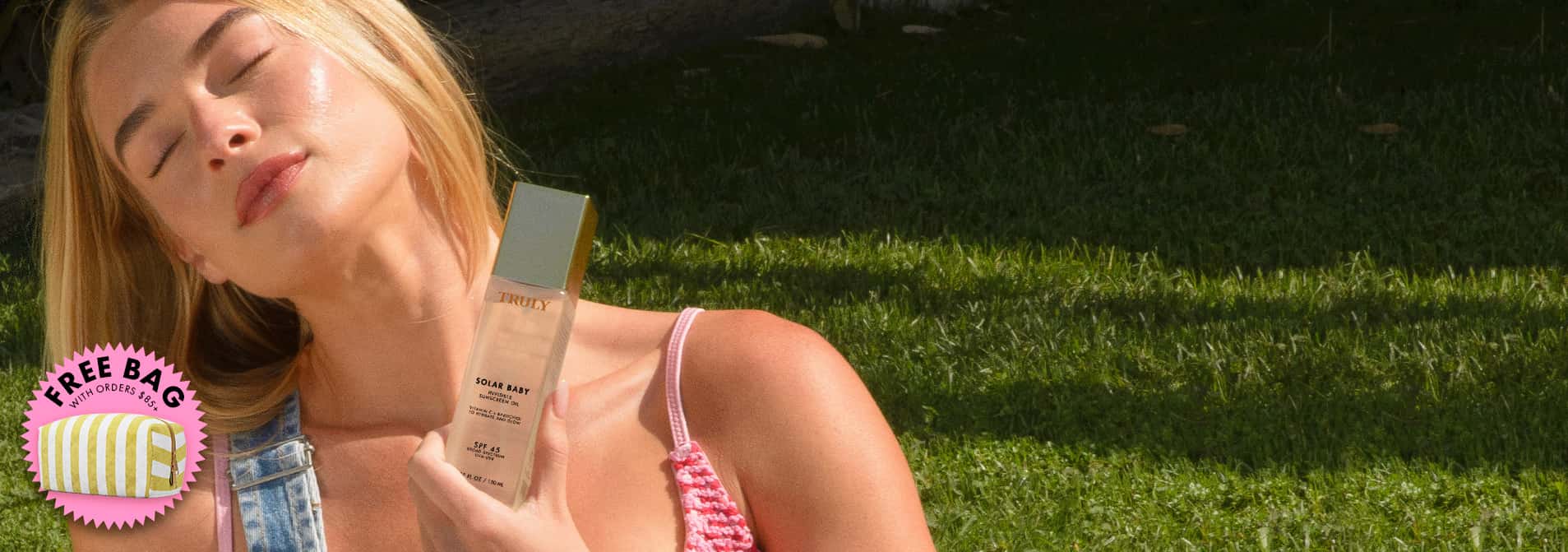 Woman lying on grass holding a bottle of Solar Baby Invisible Sunscreen Oil.
