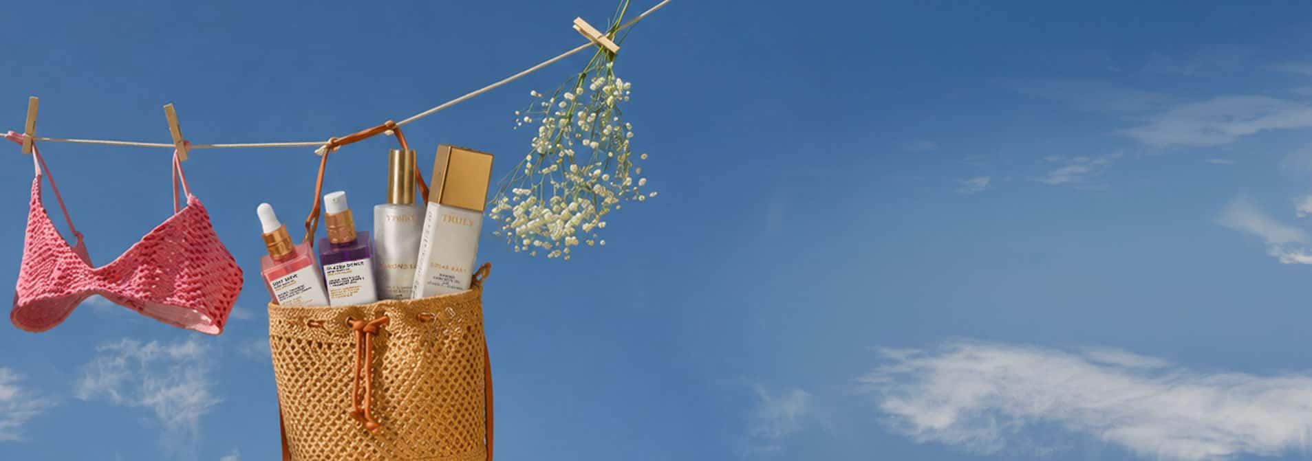 A straw tote bag hanging outdoors against a blue sky, filled with sunscreen and skincare bottles, with a pink bikini top and small white flowers clipped to a clothesline nearby.