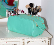 A turquoise Truly Beauty Signature Vanity Case made from vegan leather with gold zippers, embossed with TRULY, rests on a white vanity near makeup brushes and grooming items, with part of a mirror visible in the background.
