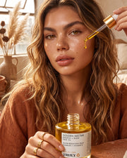 A woman with wavy hair applies Truly Beauty’s Golden Nectar Nourish + Glow Face Oil, infused with vitamin E, from a dropper bottle in a sunlit room adorned with earthy tones and dried plants, enhancing her radiant complexion.