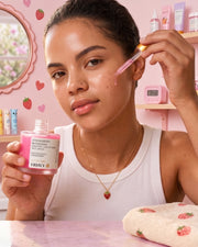 A woman applies Truly Beauty’s Strawberry Blossoms Brighten + Retexturize Face Serum—formulated with niacinamide and strawberry extract—to her cheek using a dropper, holding the bottle in a pink-themed bathroom surrounded by skincare products, making it an ideal choice for those wanting to brighten their skin.