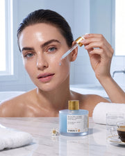 A woman applies Truly Beauty’s Glacial Glaze Hydrate + Replenish Face Serum to her cheek with a dropper in the bathroom. She holds the product’s jar, recognized for boosting skin elasticity, while a glass of water and a rolled towel sit on the counter nearby.