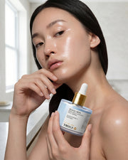 A woman with glowing, dewy skin holds a jar of Truly Beauty Glass Skin Brighten + Smooth Face Serum, gently touching her chin and looking at the camera in a softly lit bathroom setting. The hydrating serum from Truly Beauty enhances her radiant complexion, giving her that coveted glass skin glow.