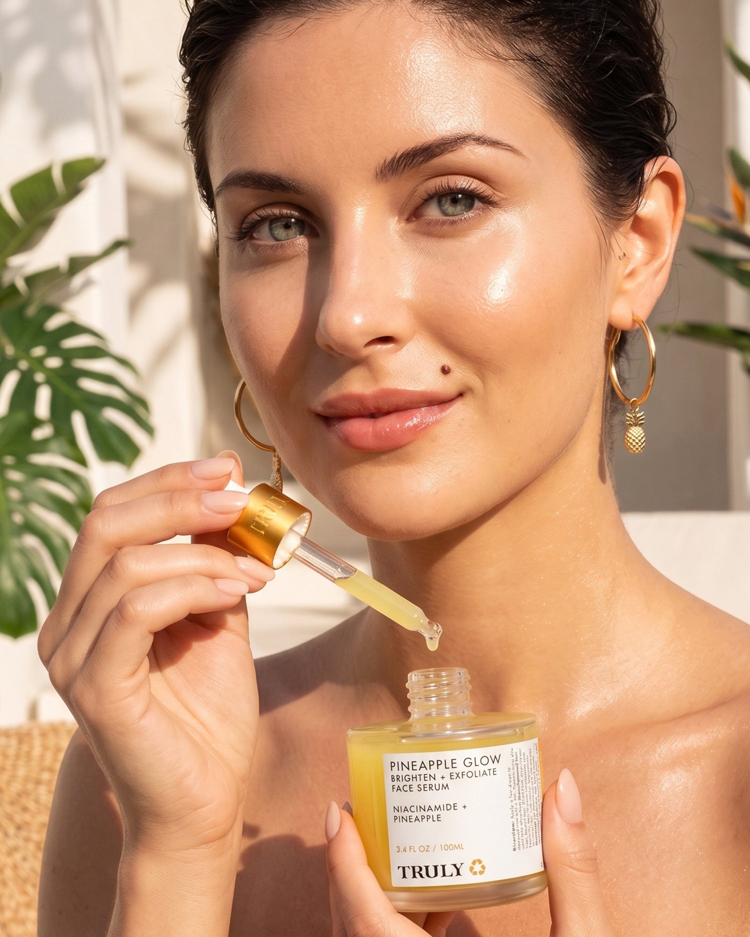 A woman with glowing skin holds a dropper and a jar labeled Pineapple Glow Brighten + Exfoliate Face Serum from Truly Beauty, celebrated for its potent Pineapple Extract that refines pores and gently exfoliates the skin. She is about to apply the serum to her face, surrounded by lush tropical plants in the background.