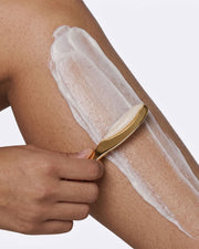 A hand uses a gold spatula to spread Truly Beauty’s Coco Cloud Hair Removal Cream onto a person’s lower leg against a plain background.