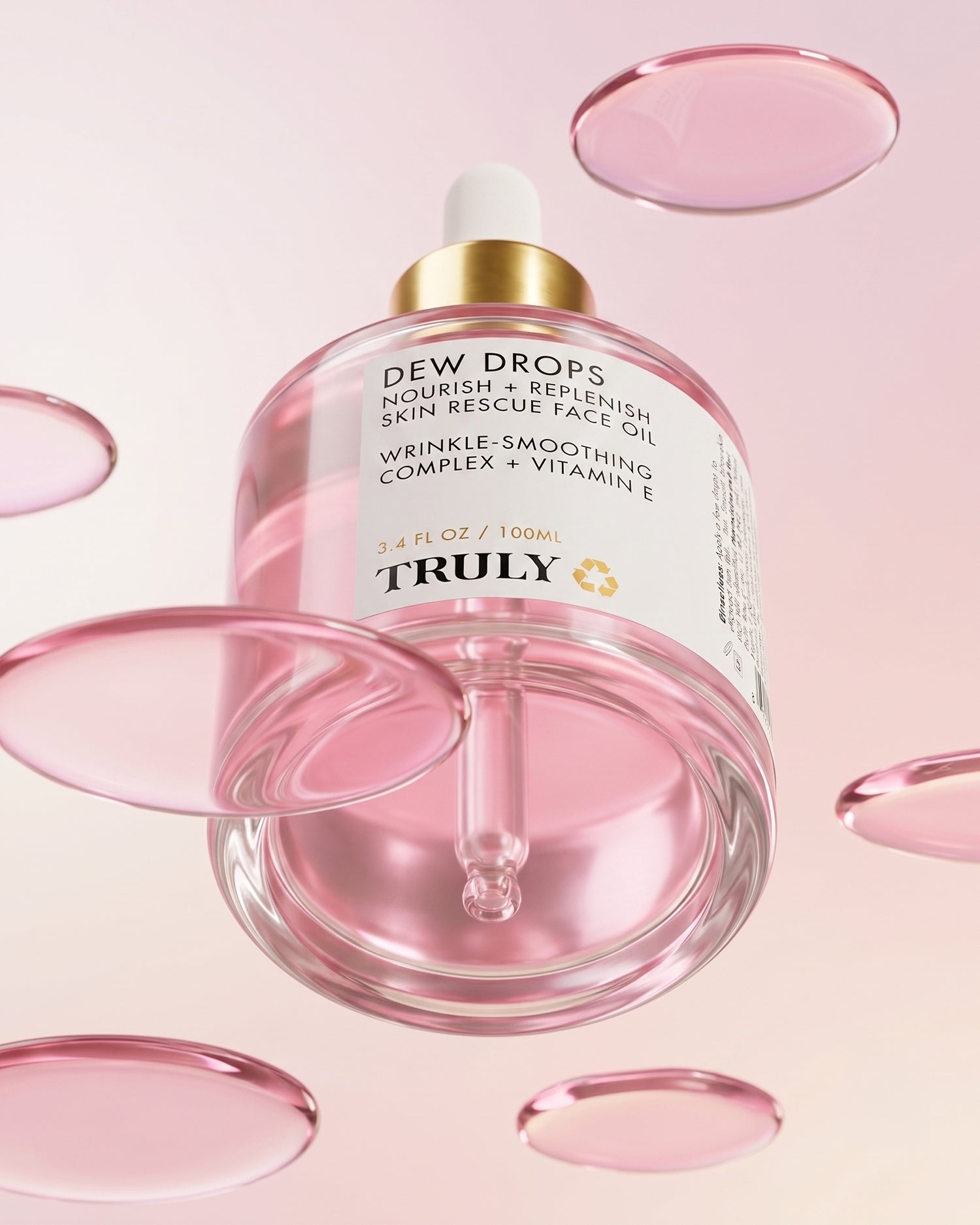 A glass dropper bottle labeled Pink Dew Drop Skin Rescue Face Oil by Truly Beauty floats among translucent pink droplets against a soft pink background. Infused with Vitamin E for wrinkle smoothing benefits, this luxurious facial oil features a gold cap with a white dropper top.