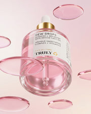 A glass dropper bottle labeled Pink Dew Drop Skin Rescue Face Oil by Truly Beauty floats among translucent pink droplets against a soft pink background. Infused with Vitamin E for wrinkle smoothing benefits, this luxurious facial oil features a gold cap with a white dropper top.