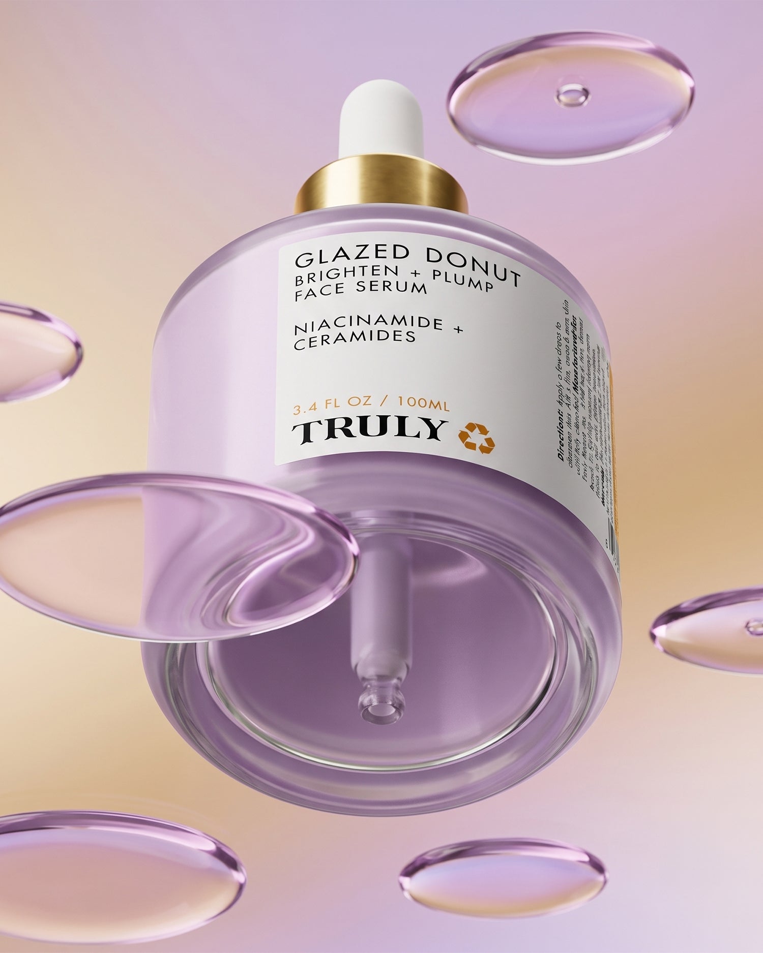 A bottle of Truly Beauty Glazed Donut Brighten + Plump Face Serum, complete with a dropper, floats among translucent liquid droplets against a pastel backdrop. Ideal for achieving luminous Glazed Donut Skin, this hydrating serum’s label spotlights niacinamide and ceramides.