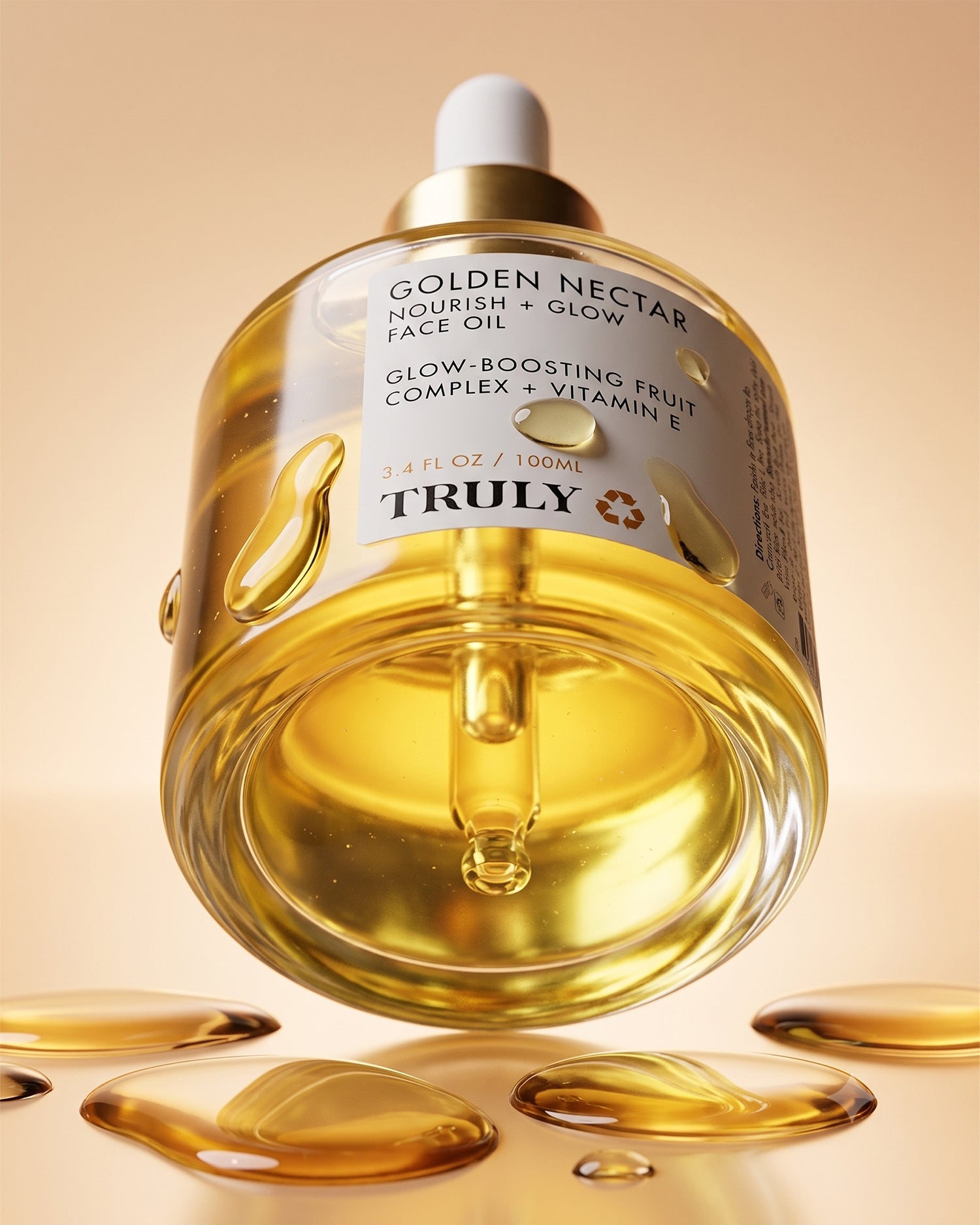A close-up of a glass bottle of Truly Beauty Golden Nectar Nourish + Glow Face Oil with a dropper, golden liquid, and oil droplets resting on a reflective surface against a light golden background, highlighting its infusion of vitamin E for radiant, glowing skin.