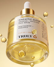 A glass bottle of Truly Beauty Pineapple Glow Brighten + Exfoliate Face Serum, featuring pineapple extract and a white dropper cap, is surrounded by floating serum droplets on a yellow gradient background, formulated to exfoliate and refine pores.