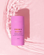 A Unicorn Fruit Skin Brightening and Smoothing Deodorant stick from Truly Beauty sits on a pink surface with water ripples. This vegan, aluminum-free deodorant brightens and smooths your underarms for elevated care.