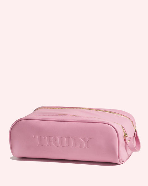 The Truly Beauty Signature Vanity Case is a pink rectangular pouch made from vegan leather, featuring gold zippers and TRULY embossed on the front, shown against a light pink background.