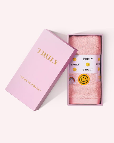 A pink box labeled Truly Beauty contains a folded Pour le Visage Face Towel with a pink wrap featuring smiley faces, rainbows, and TRULY in black. The box lid sits to the left of the open box.