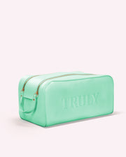 The Signature Vanity Case by Truly Beauty is a mint green rectangular vegan leather bag with gold zippers and handles, TRULY embossed on the side—perfect as a Skincare Travel Pouch, shown on a pale pink background.