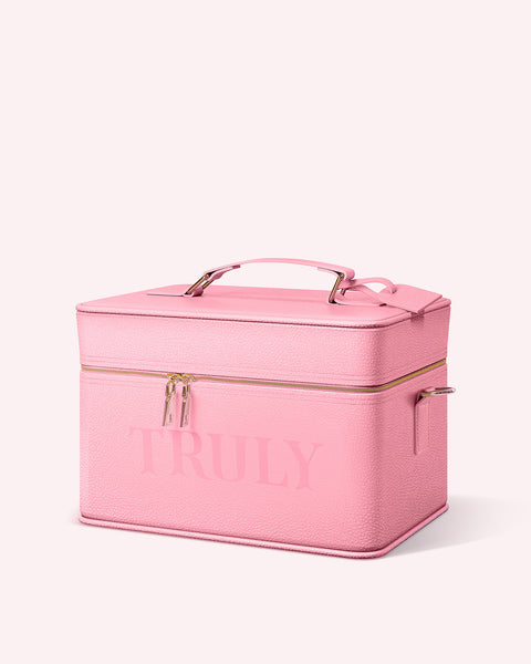 The Truly Beauty Truly Signature Travel Trunk is a rectangular, textured pink case with a top handle, gold zippers, and TRULY on the front—ideal for skincare or as an overnight trunk. Background is light pink.