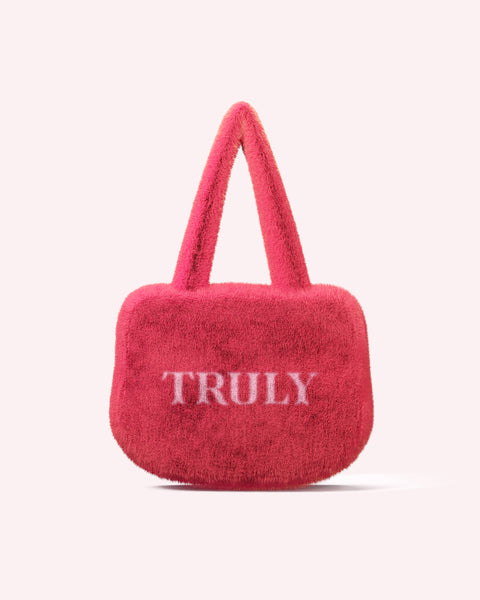 The Pink Fuzzy Bag by Truly Beauty is a vibrant pink clutch with two handles and TRULY in white letters on branded fabric, shown against a pale pink background.
