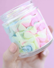 A hand with light pink nail polish holds a clear jar filled with pastel-colored marshmallows against a pink background, echoing the whimsical colors of Truly Beauty’s Unicorn CBD Soothe & Glow Whipped Body Butter—an ideal treat for calming inflammation and indulging your senses.
