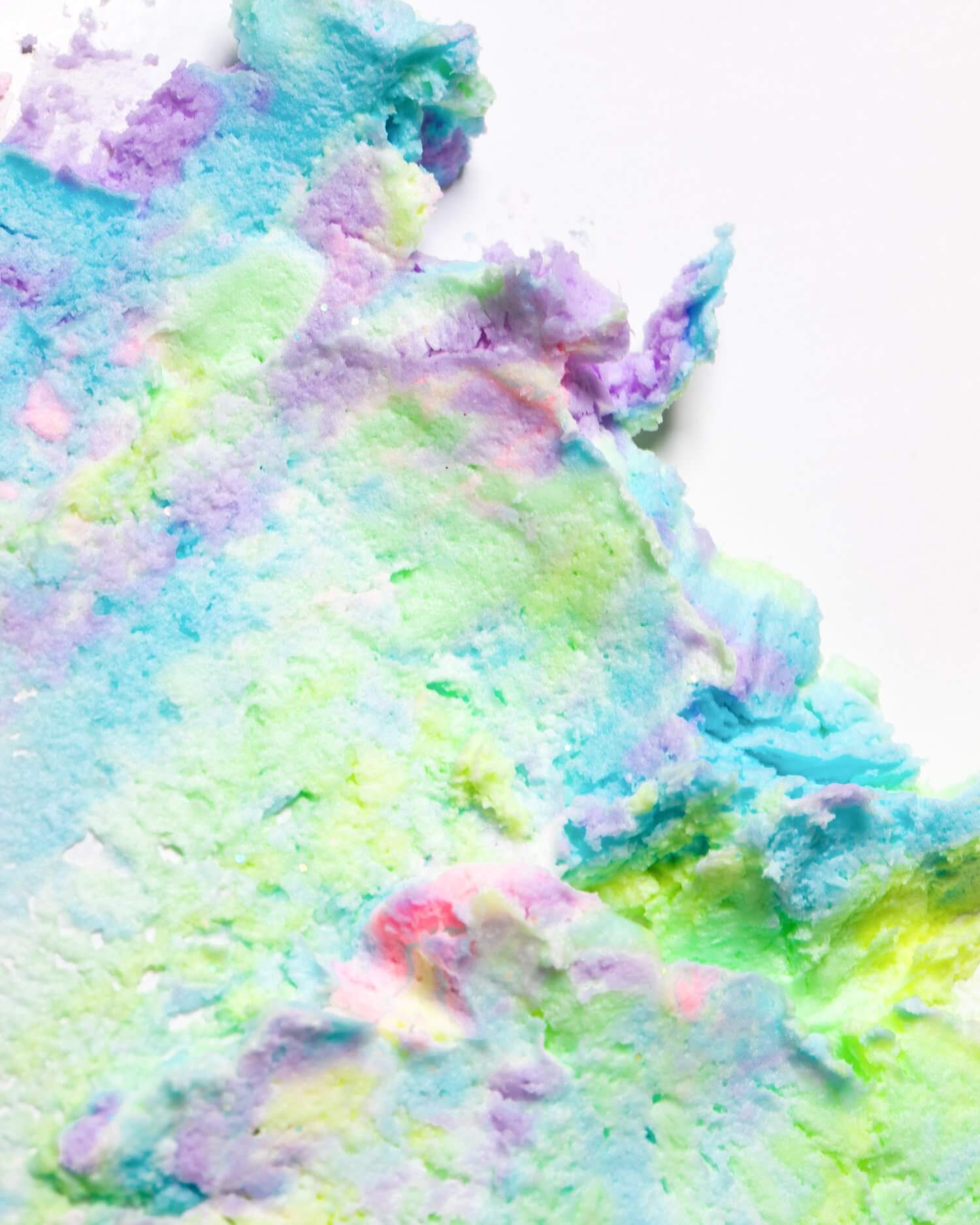 A vibrant, pastel-textured swirl spread across a white surface—showcasing shades of blue, green, purple, and touches of pink and yellow—reminiscent of whipped or spread clay or dough. This dreamy composition evokes the look of Truly Beauty’s Unicorn CBD Soothe & Glow Whipped Body Butter, crafted to calm inflammation.