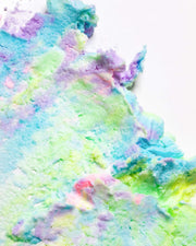 A vibrant, pastel-textured swirl spread across a white surface—showcasing shades of blue, green, purple, and touches of pink and yellow—reminiscent of whipped or spread clay or dough. This dreamy composition evokes the look of Truly Beauty’s Unicorn CBD Soothe & Glow Whipped Body Butter, crafted to calm inflammation.